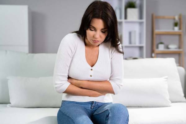 A woman sitting on a couch, clutching her stomach in discomfort, experiencing symptoms of diarrhea or a urinary infection.