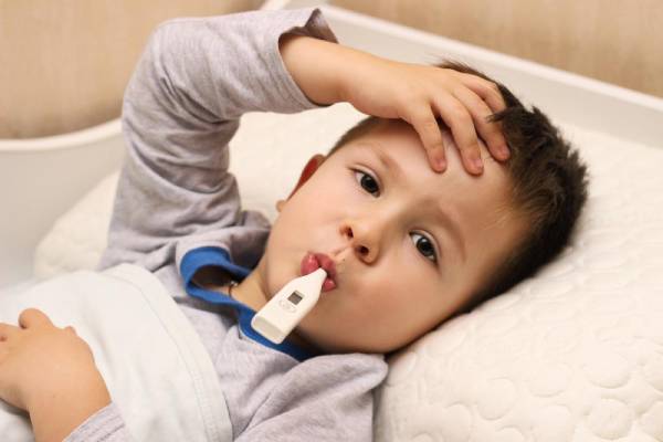 A sick child lying in bed with a thermometer in his mouth, showing signs of fever and discomfort.